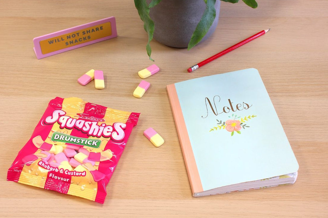 Squashies Drumsticks candy package, notebook, and candy on a wooden table with a plant in the background.