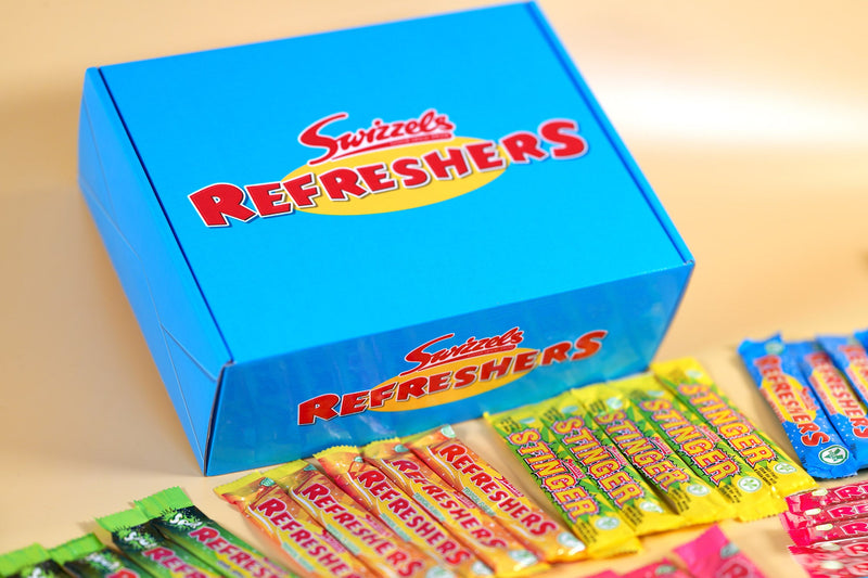 A bright blue Swizzels Refreshers box with the brand logo in red and yellow, shown with several individually wrapped Refreshers candy bars in front on a warm beige background.