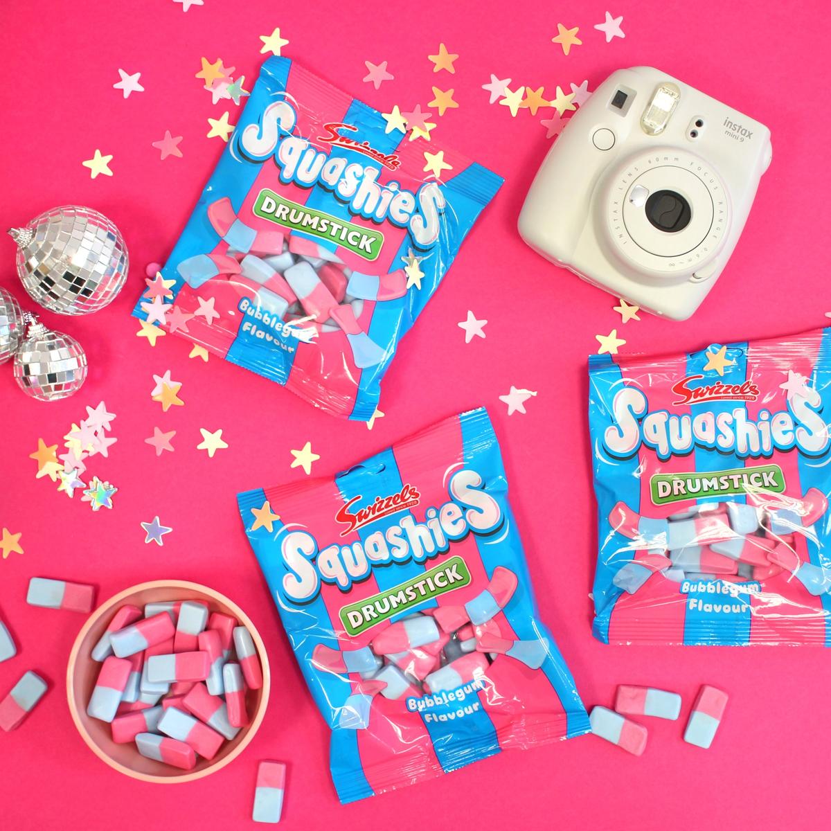 Squashies Drumstick candy packages on a pink background with a camera and confetti.