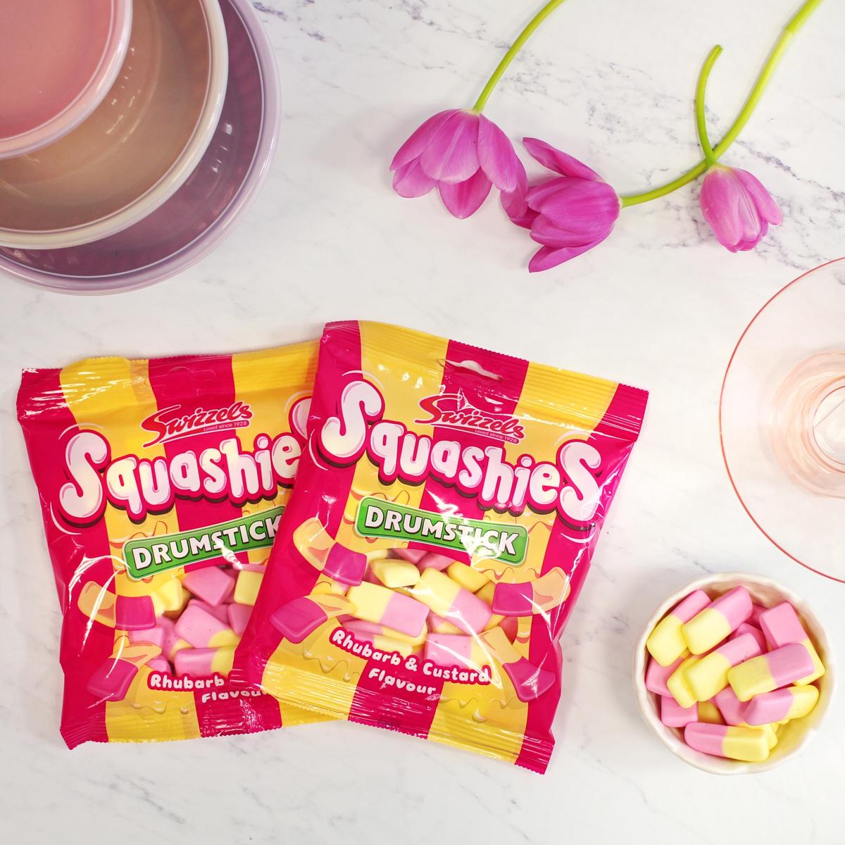 Two packets of Swizzles Squashies Drumstick on a white surface with pink flowers and a cup.
