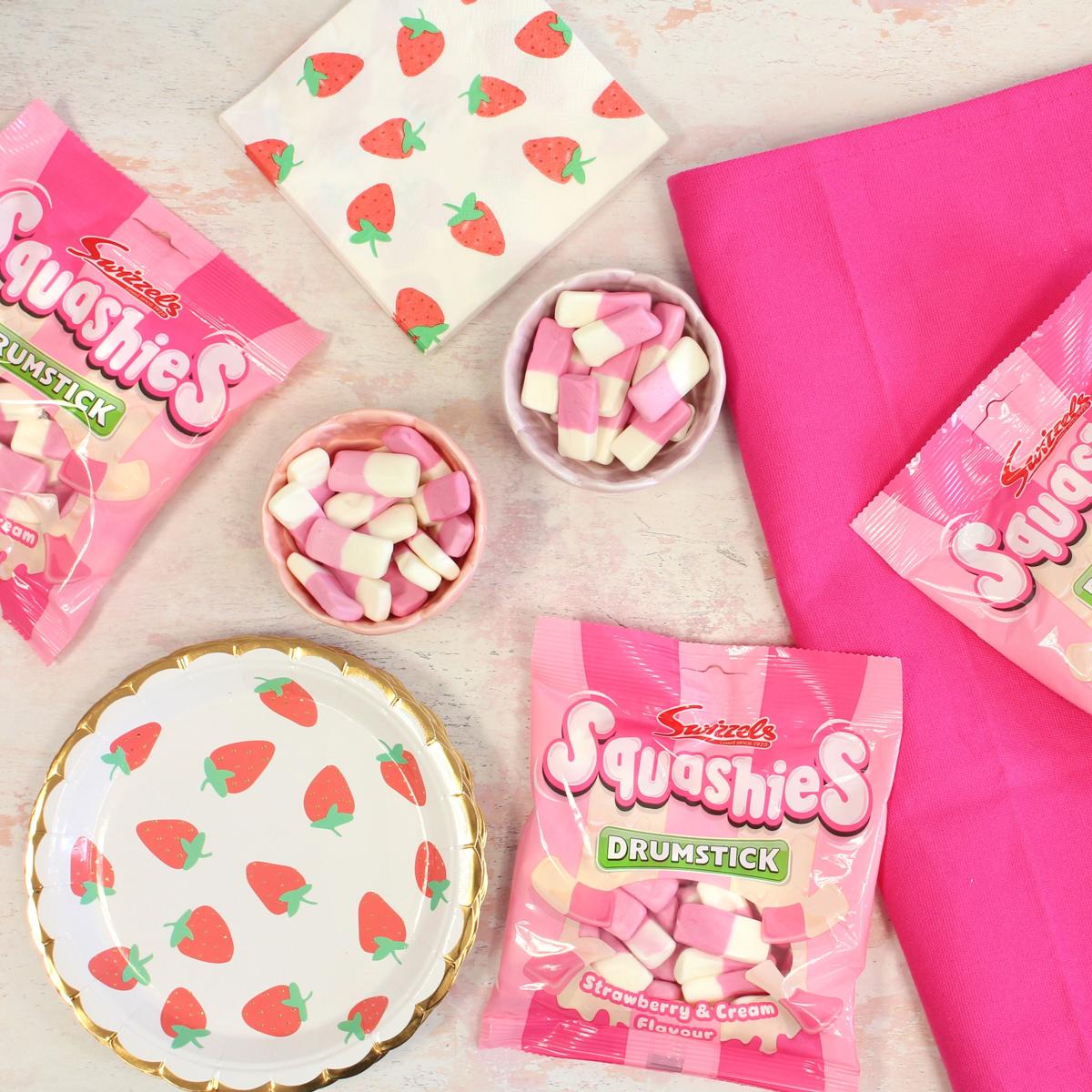 Squashies Strawberry & Cream flavor candy packages with pink and white paper plates and napkins on a light background.