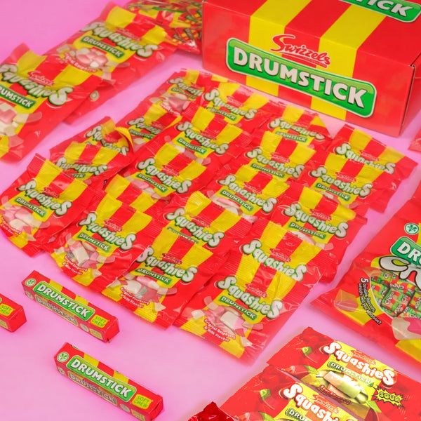 A collection of Snickers Drummick Sticks and Sticks Coos candy products arranged on a solid pink background, including a large red and yellow box, individual stick packs, and wrapped candy pieces.