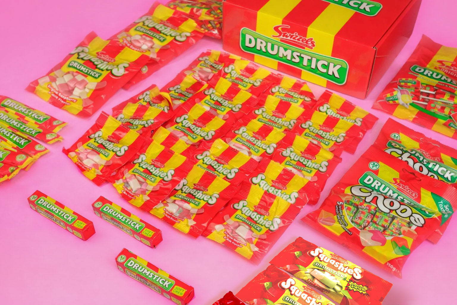 A collection of Snickers Drummick Sticks and Sticks Coos candy products arranged on a solid pink background, including a large red and yellow box, individual stick packs, and wrapped candy pieces.
