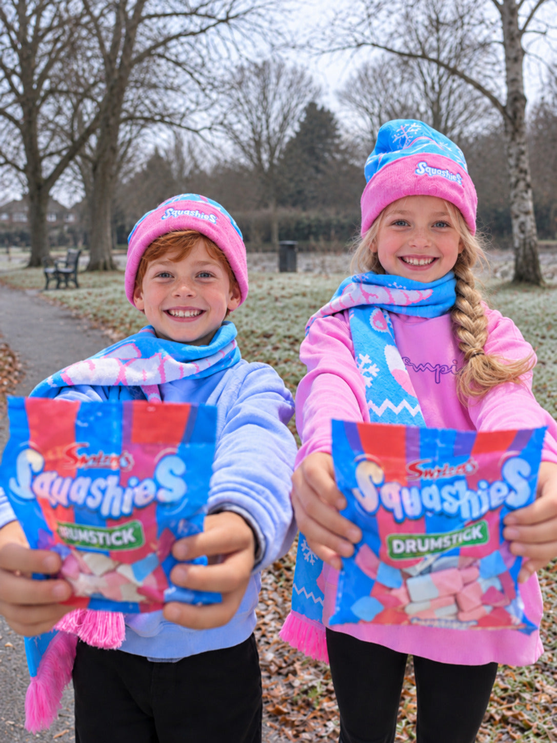 Two children outdoors in Squashies themed blue and pink winter clothing, each holding up a bag of Squashies Drumstick sweets toward the camera.