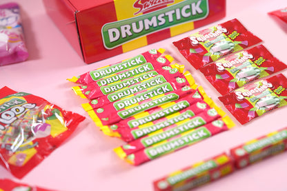 A close-up, angled view of a Sweets by Dr. Drumstick candy assortment, showing multiple individually wrapped red and pink candy sticks and a larger red box on a solid pink background.