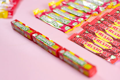 Several packs of Drummstick Refreshers gum are arranged on a soft pink background, with some packs in sharp focus and others blurred in the background.