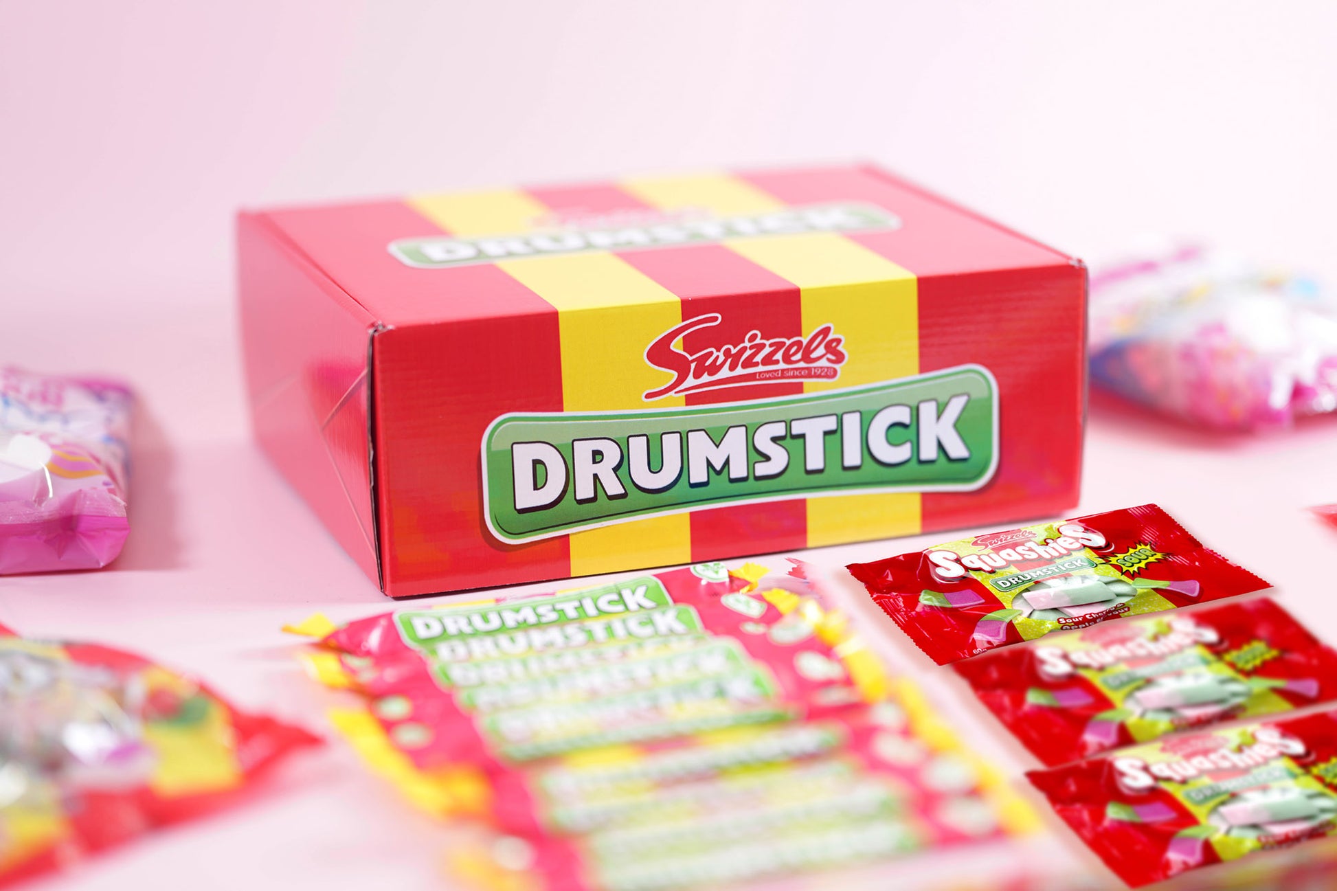 A box of Swizzels Drumstick candy on a light pink background, with several individually wrapped drumsticks arranged in front.