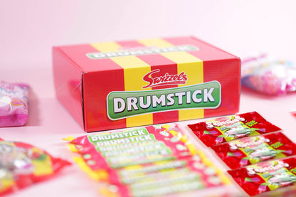 A box of Swizzels Drumstick candy on a light pink background, with several individually wrapped drumsticks arranged in front.