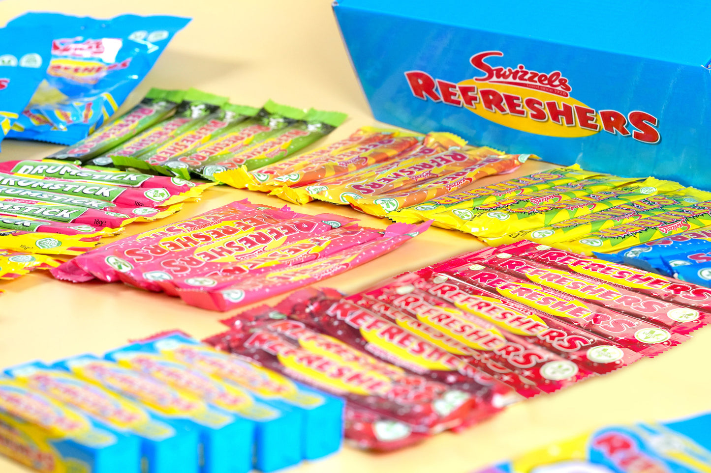 A variety of Swizzels Refreshers candy sticks and packs arranged on a light beige surface, with a blue box of Swizzels Refreshers in the background.