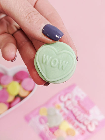 Hand holding a green heart-shaped candy with 'WOW' text against a pink background