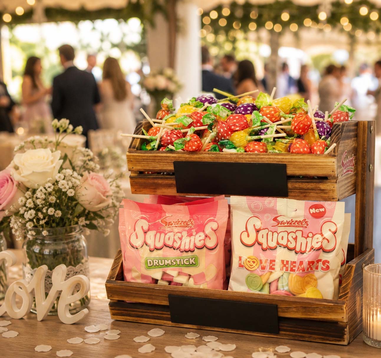 Sweet treats including Squashies candy packages and fruit skewers on a wooden tray with blurred people in the background.
